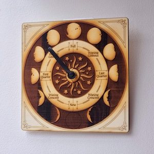 May include: A wooden moon phase clock with a black hand pointing to the full moon. The clock face is decorated with a sun and stars. The text on the clock face reads "Full", "Waning Gibbous", "Last Quarter", "Waning Crescent", "New", "Waxing Crescent", "First Quarter", and "Waxing Gibbous".