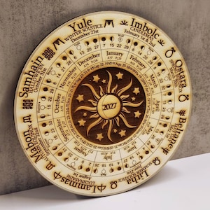 May include: A circular wooden calendar for 2027 featuring laser-engraved text and graphics. The design includes the months of the year, moon phases, zodiac signs, and seasonal festivals like Yule, Imbolc, Ostara, Beltane, Litha, Lammas, Mabon, and Samhain.