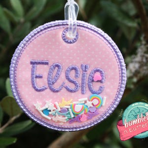 May include: A pink, round, embroidered tag with the name "Elsie" in purple script. The tag has a purple border and is filled with colourful confetti and a clear loop for hanging. The tag is decorated with a pink heart.