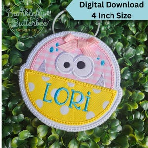 May include: A round embroidered patch featuring a pink and white gingham monster with two large eyes, a light pink ribbon bow, and a yellow bottom section with white polka dots. The name LORI is stitched in blue. It measures 4 inches in size.