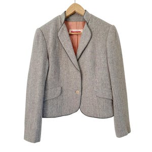 May include: A light gray blazer with a single button closure and two front pockets. The blazer has a black trim and a peach-colored lining. The label "foxanne" is visible on the inside. The jacket is hanging on a wooden hanger.