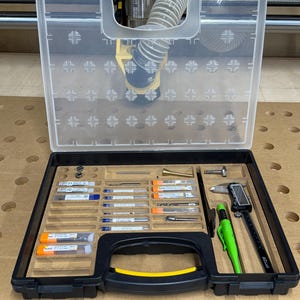 Pode incluir: Uma caixa de plástico preta com uma alça amarela, contendo uma variedade de ferramentas pequenas, incluindo um lápis verde, um paquímetro prateado e vários tubos pequenos e transparentes com pontas metálicas dentro. A caixa está aberta e está sobre uma superfície de madeira.