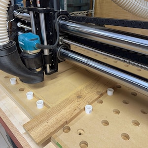 Könnte beinhalten: Nahaufnahme einer CNC-Maschine mit einem Fräser. Die Maschine ist auf einer Arbeitsfläche mit einem Stück Holz aufgebaut, das geschnitten wird. Die Maschine hat ein Staubabsaugsystem.