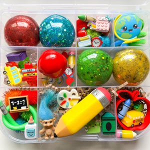 May include: A clear plastic organizer box filled with school-themed sensory items including red and green glitter spheres, a globe, a yellow pencil-shaped eraser, a small troll doll, miniature school buses, crayons, and a chalkboard with 1-3=? written on it.