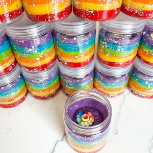 May include: Clear plastic jars filled with colorful rainbow-layered slime. Each jar has a clear lid. One jar is open, revealing a purple top layer with a flower-shaped charm. Glitter accents the layers. A fun, sensory toy.