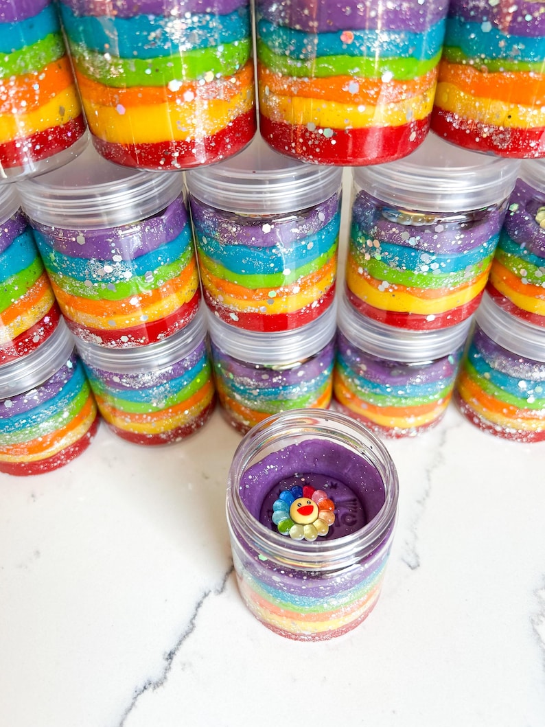 May include: Clear plastic jars filled with colorful rainbow-layered slime. Each jar has a clear lid. One jar is open, revealing a purple top layer with a flower-shaped charm. Glitter accents the layers. A fun, sensory toy.