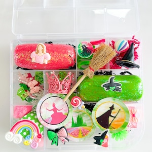 May include: A clear plastic organizer filled with various themed accessories. Includes a pink princess-themed item, a green witch-themed item, a rainbow, a crown, bows, and a miniature broom. The organizer is divided into multiple compartments.