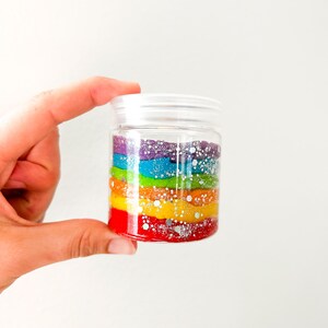 May include: A clear plastic jar filled with layers of colorful, glittery material in a rainbow pattern. The colors include red, orange, yellow, green, blue, and purple. The jar has a white lid and is held by a hand.