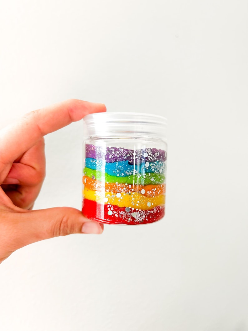 May include: A clear plastic jar filled with layers of colorful, glittery material in a rainbow pattern. The colors include red, orange, yellow, green, blue, and purple. The jar has a white lid and is held by a hand.