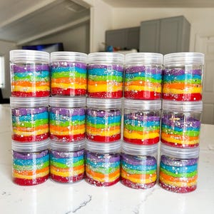 May include: A collection of clear plastic jars filled with colorful, layered rainbow-colored material. Each jar has a clear lid and contains layers of red, orange, yellow, green, blue, and purple. Silver glitter is sprinkled between the layers.