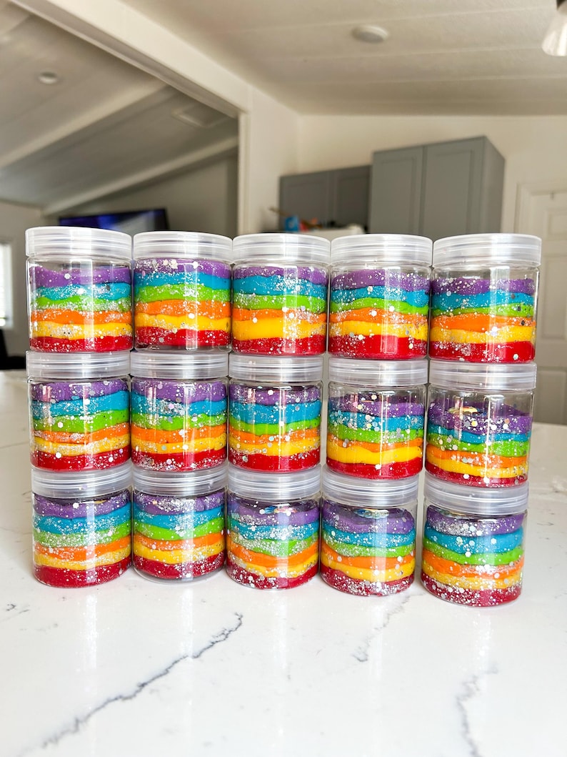 May include: A collection of clear plastic jars filled with colorful, layered rainbow-colored material. Each jar has a clear lid and contains layers of red, orange, yellow, green, blue, and purple. Silver glitter is sprinkled between the layers.