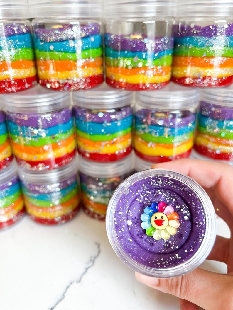 May include: Clear plastic jars filled with rainbow-colored layers of what appears to be slime or play dough. Each jar has a silver lid. One jar is open, revealing purple slime with a colorful flower decoration.