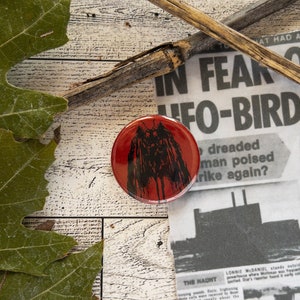 May include: A red button with a black and white image of a moth with large wings. The button is on a wooden surface with a newspaper in the background. The newspaper headline reads "In Fear of UFO-Bird".