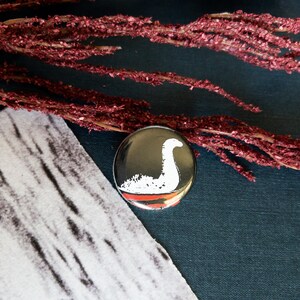 May include: A round, silver-colored button featuring a white illustration of the Loch Ness Monster against a black background. A red stripe is at the bottom. The button is placed on a dark blue surface, with a piece of patterned fabric and red dried flowers.