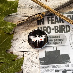 May include: A black button with a white moth with red eyes on it. The button is on top of a newspaper with the headline "In Fear of UFO-Bird".