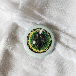 May include: A round button with a green and yellow eye design. The eye has a black pupil and a white highlight. The button is on a white fabric background.