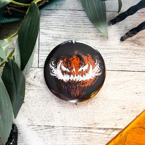 May include: A black and orange round button with a white scary pumpkin face graphic. The button is on a white wooden surface.