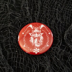 May include: A red button with a white image of a devil with horns and a tongue sticking out. The devil is surrounded by a white chain link design.