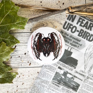 May include: A white round button featuring a black and red illustration of a winged creature with glowing red eyes. The button is placed on a wooden surface with a newspaper clipping that reads "IN FEAR UFO-BIRD".