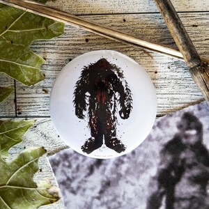 May include: A white button with a black and red silhouette of Bigfoot, a mythical creature, standing in the center. The button has a white background and a black border.
