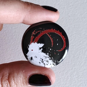 May include: A black and white button with a red design. The design features a white goat with a red horn and a black background with white splatters.