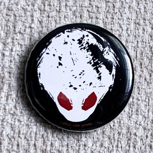 May include: Black and white button with a white alien head with red eyes. The text "other side prints" is on the bottom of the button.