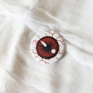 May include: A round button with a red eye and blood vessels on a white background. The eye is black with a white pupil.