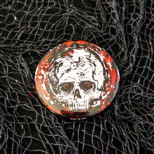 May include: A round button with a black, white, and red design. The design features a skull with red splatters of paint. The text "Wicker did it first" is printed on the bottom of the button.