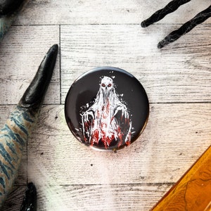 May include: A round, black button featuring a white ghost-like figure with red accents and eyes. The button has a spooky, Halloween theme. The background is a light wood surface with decorative elements.