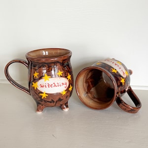 Könnte beinhalten: Zwei braune Keramikbecher mit Henkeln und kleinen Füßen. Jeder Becher hat das Wort "witchling" in gelber Schrift auf einem weißen Oval, umgeben von gelben Sternen. Ein Becher steht aufrecht, der andere liegt auf der Seite.
