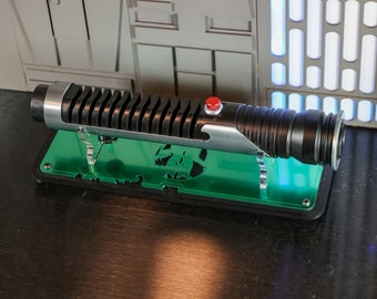 Op de afbeelding: Een zwart lightsaber met een zilveren handvat en een rode knop wordt getoond op een groene standaard. De standaard heeft een uitgesneden ontwerp.