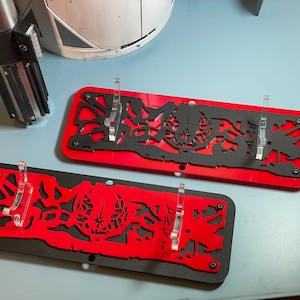 May include: Two black and red lightsaber display stands with clear acrylic holders. The stands feature a detailed design of a lightsaber blade and a circular pattern.