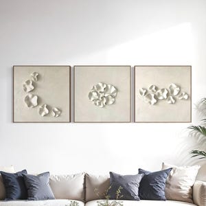 May include: Three square wall art pieces with white, abstract floral designs in wooden frames. The artwork hangs above a cream-colored sofa with navy blue throw pillows. A wooden coffee table and a potted plant complete the living room setting.