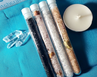 May include: Four glass tubes filled with different colored bath salts. The salts are pink, white, black, and beige. The tubes are on a blue surface with two clear quartz crystals.