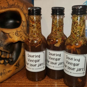 May include: Three small glass bottles filled with a brown liquid and herbs. Each bottle has a white label that reads "Souring Vinegar for sour jars!"