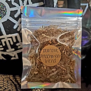 May include: A clear, iridescent zip-lock bag filled with dried herbs. A small, round, brown sticker on the bag reads "Remove attachment blend" in handwritten style. The background includes a black surface with white symbols and a decorative item.