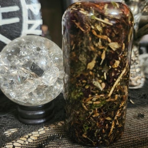 May include: A clear glass bottle filled with a mixture of dried herbs and flowers. The bottle is sitting on a black surface with a crystal ball in the background.