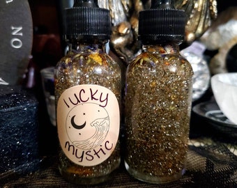 May include: Two glass bottles filled with a shimmering, golden liquid. Each bottle has a black dropper top and a label that reads "lucky mystic" with a crescent moon and wave design. The liquid contains small particles and visible botanicals.