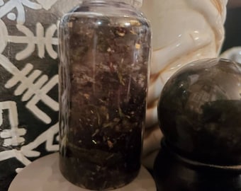 May include: A clear glass bottle filled with a dark brown liquid and various herbs and plant matter. The bottle is sitting on a white surface.