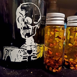May include: Two small glass bottles with silver lids filled with a yellow liquid and red pepper flakes. The bottles are sitting on a black surface in front of a black mug with a white skull and skeleton design.