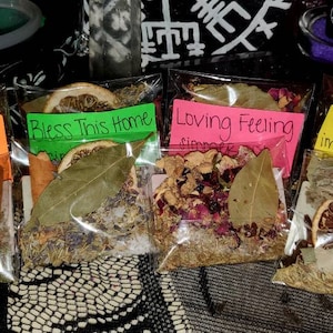 May include: Four small clear plastic bags filled with dried herbs, spices, and fruit peels. The bags are labeled with handwritten text: "Money Magnet", "Bless This Home", "Loving Feeling", and "4 Thieves Immune Boosting".