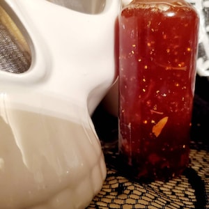 Pu&ograve; includere: Una bottiglia di vetro trasparente riempita di un liquido rosso con glitter dorati. La bottiglia &egrave; posizionata accanto a un teschio di ceramica bianca.