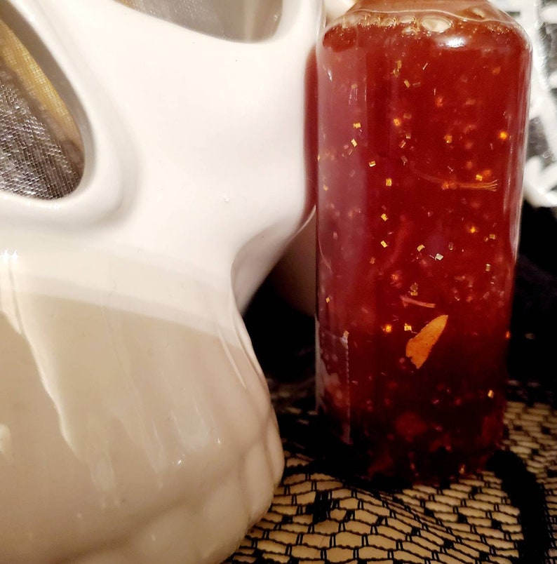 Pu&ograve; includere: Una bottiglia di vetro trasparente riempita di un liquido rosso con glitter dorati. La bottiglia &egrave; posizionata accanto a un teschio di ceramica bianca.