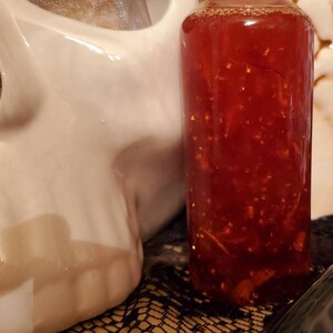 Pu&ograve; includere: Una bottiglia di vetro piena di un liquido rosso contenente glitter dorati. La bottiglia &egrave; appoggiata su una superficie con un motivo bianco e nero.