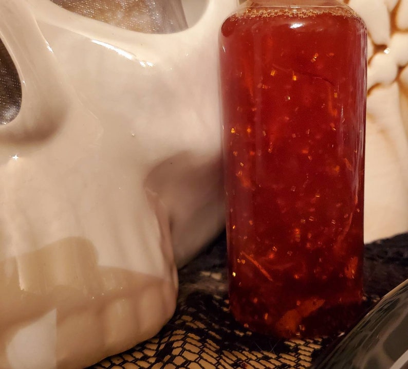 Pu&ograve; includere: Una bottiglia di vetro piena di un liquido rosso contenente glitter dorati. La bottiglia &egrave; appoggiata su una superficie con un motivo bianco e nero.