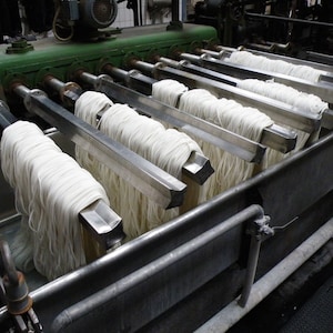 Puede incluir: Primer plano de una máquina utilizada para procesar hilo. La máquina tiene múltiples varillas de metal con hilo blanco colgando de ellas. El hilo está sumergido en una gran tina de metal.