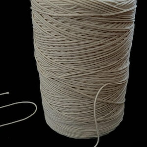 May include: A spool of natural colored cotton cord, wound tightly on a cardboard tube. The cord is smooth and has a subtle sheen. A length of cord is hanging off the spool.