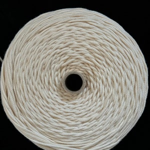 Könnte beinhalten: Nahaufnahme einer Spule cremefarbener Wolle, eng in konzentrischen Kreisen gewickelt. Die Wolle hat eine sichtbare Textur und ein zentrales Loch. Der Hintergrund ist schwarz, wodurch die Form und Textur der Wolle hervorgehoben werden, ideal für Bastelarbeiten.
