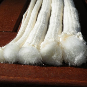May include: Close-up of five white cotton cords with fluffy ends. The cords are arranged in a row on a brown wooden surface.
