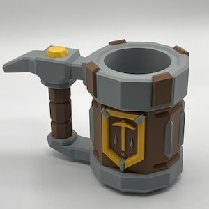 May include: A gray, brown, and yellow tankard with a faceted, geometric design. The handle is shaped like a hammer with a yellow top accent. A central shield emblem features a yellow border and a pickaxe icon. A US quarter coin sits nearby.